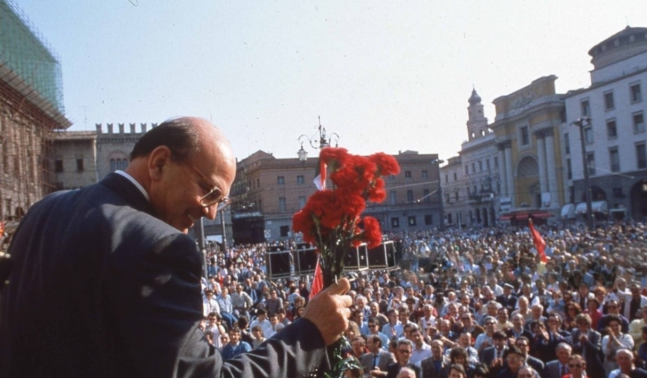 Bettino Craxi, politico con garofani rossi saluta una folla in una piazza storica durante un evento pubblico sotto il sole.