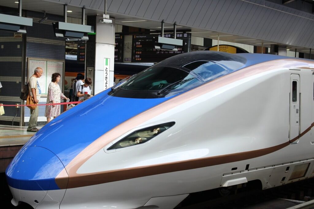 Treno ad alta velocità bianco e blu fermo in stazione, con passeggeri in attesa sulla banchina e tabelloni delle partenze sullo sfondo.