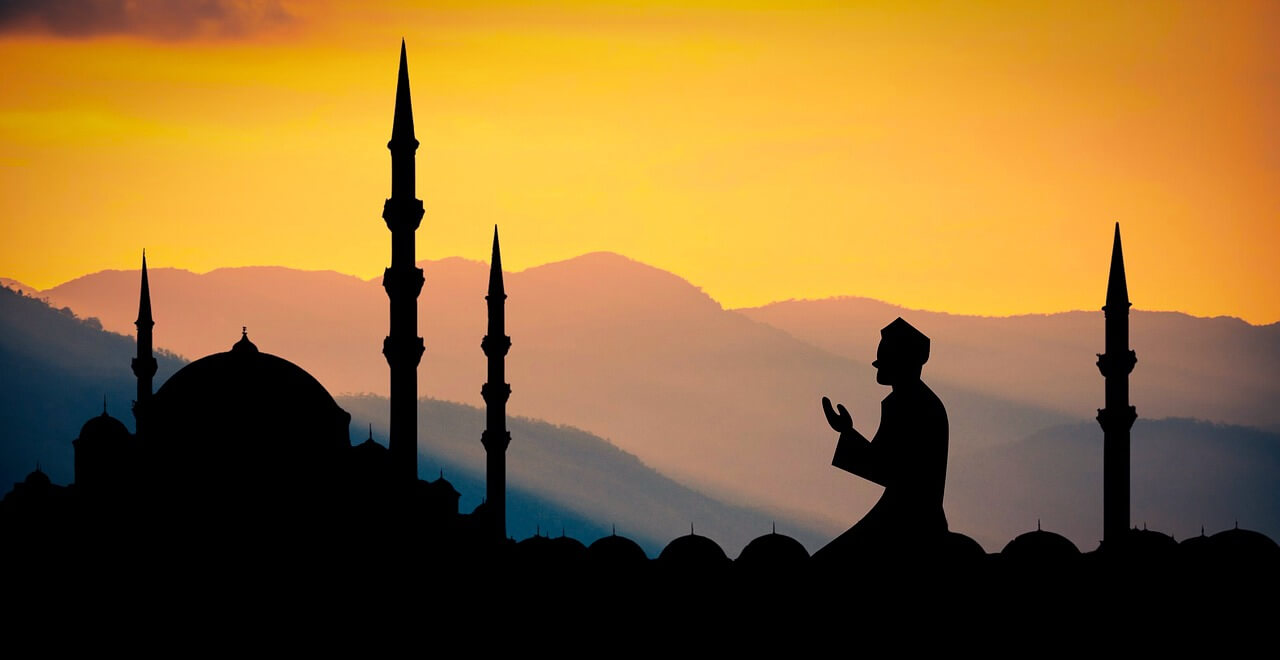 Silhouette di una moschea con minareti e cupole, con una figura di una persona che prega di fronte a un cielo arancione e montagne in lontananza.