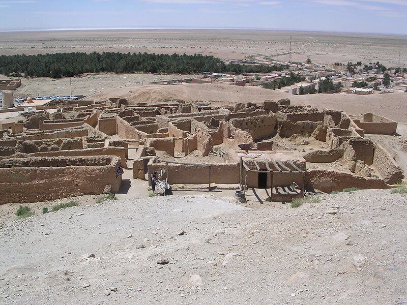 Vista panoramica delle rovine di un insediamento fortificato in argilla, con mura parzialmente crollate, situate in un ambiente desertico con una lussureggiante oasi di palme visibile in lontananza.