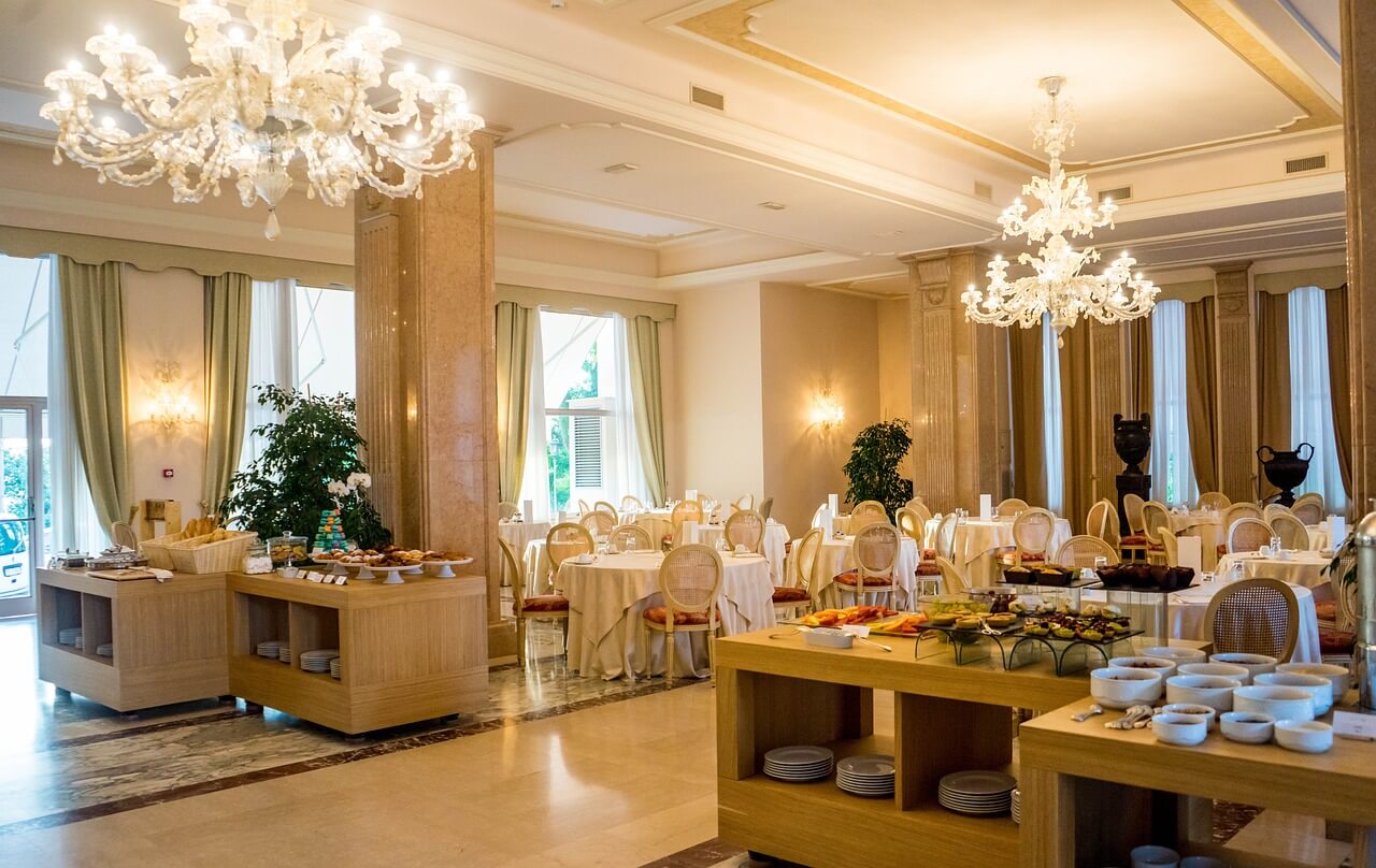 Raffinata sala da pranzo di un hotel di lusso con tavoli eleganti, lampadari di cristallo e buffet ricco di pietanze. Ambiente luminoso e accogliente.