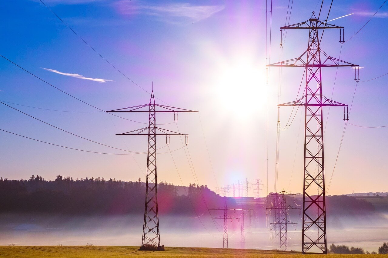 High-voltage electricity pylons with power lines stretch across a foggy landscape at sunrise, with the sun casting a golden glow over the scene.