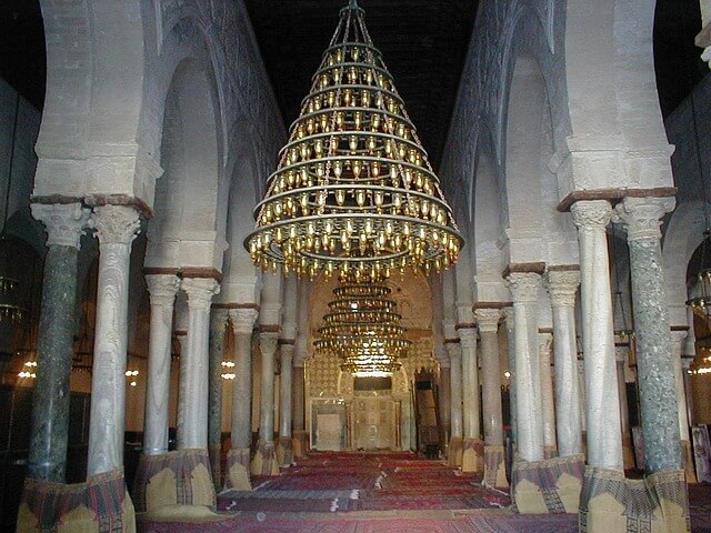 Interno della Moschea di Kairouan in Tunisia, con colonne in marmo, grandi lampadari dorati e tappeti rossi sul pavimento.